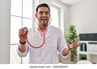 Young hispanic doctor man with beard holding stethoscope auscultating celebrating crazy and amazed for success with open eyes screaming excited.  - Shutterstock ID 2205185419