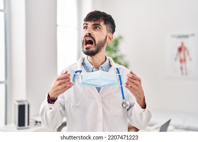 Young hispanic doctor man with beard holding safety mask angry and mad screaming frustrated and furious, shouting with anger. rage and aggressive concept.  - Shutterstock ID 2207471223