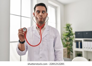 Young hispanic doctor man with beard holding stethoscope auscultating making fish face with mouth and squinting eyes, crazy and comical.  - Shutterstock ID 2281212281