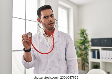 Young hispanic doctor man with beard holding stethoscope auscultating in shock face, looking skeptical and sarcastic, surprised with open mouth  - Shutterstock ID 2306087935