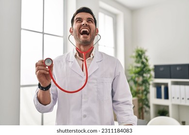 Young hispanic doctor man with beard holding stethoscope auscultating angry and mad screaming frustrated and furious, shouting with anger looking up.  - Shutterstock ID 2311184083