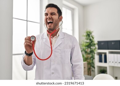 Young hispanic doctor man with beard holding stethoscope auscultating angry and mad screaming frustrated and furious, shouting with anger. rage and aggressive concept.  - Shutterstock ID 2319671413