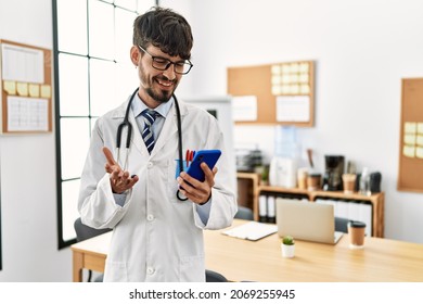 Young Hispanic Doctor Man Having Video Call Using Smartphone At Clinic