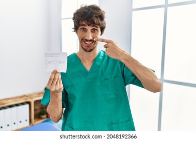 Young Hispanic Doctor Man Holding Covid Record Card Pointing With Hand Finger To Face And Nose, Smiling Cheerful. Beauty Concept 