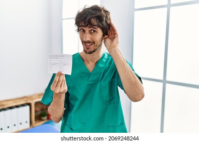 Young Hispanic Doctor Man Holding Covid Record Card Smiling With Hand Over Ear Listening An Hearing To Rumor Or Gossip. Deafness Concept. 