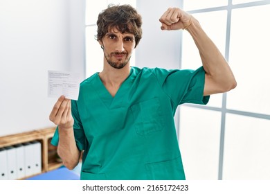 Young Hispanic Doctor Man Holding Covid Record Card Strong Person Showing Arm Muscle, Confident And Proud Of Power 