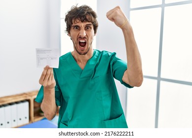 Young Hispanic Doctor Man Holding Covid Record Card Angry And Mad Raising Fist Frustrated And Furious While Shouting With Anger. Rage And Aggressive Concept. 