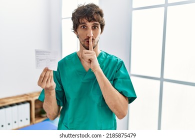 Young hispanic doctor man holding covid record card asking to be quiet with finger on lips. silence and secret concept. 