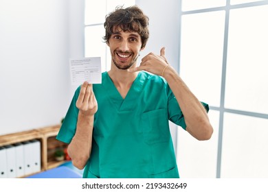 Young Hispanic Doctor Man Holding Covid Record Card Smiling Doing Phone Gesture With Hand And Fingers Like Talking On The Telephone. Communicating Concepts. 