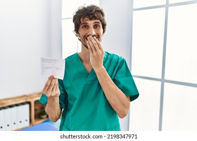 Young Hispanic Doctor Man Holding Covid Record Card Laughing And Embarrassed Giggle Covering Mouth With Hands, Gossip And Scandal Concept 