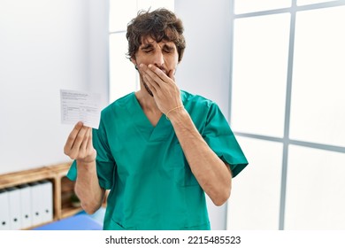 Young Hispanic Doctor Man Holding Covid Record Card Bored Yawning Tired Covering Mouth With Hand. Restless And Sleepiness. 