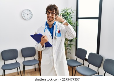 Young Hispanic Doctor Man At Waiting Room Smiling Doing Phone Gesture With Hand And Fingers Like Talking On The Telephone. Communicating Concepts. 