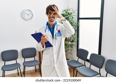 Young Hispanic Doctor Man At Waiting Room Doing Ok Gesture With Hand Smiling, Eye Looking Through Fingers With Happy Face. 