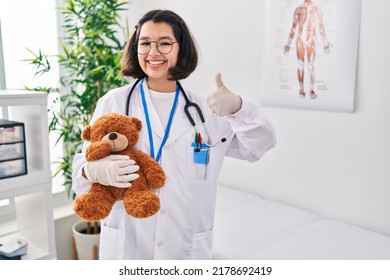 Young hispanic doctor woman holding teddy bear smiling happy and positive, thumb up doing excellent and approval sign 