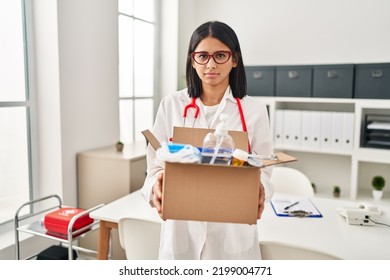 Young Hispanic Doctor Woman Holding Box With Medical Items Relaxed With Serious Expression On Face. Simple And Natural Looking At The Camera. 