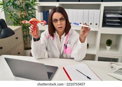 Young Hispanic Doctor Woman Holding Anatomical Model Of Female Genital Organ And Pregnancy Test In Shock Face, Looking Skeptical And Sarcastic, Surprised With Open Mouth 