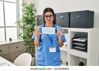 Young Hispanic Doctor Woman Holding Safety Mask At Clinic Clueless And Confused Expression. Doubt Concept. 