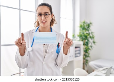 Young Hispanic Doctor Woman Holding Safety Mask Clueless And Confused Expression. Doubt Concept. 