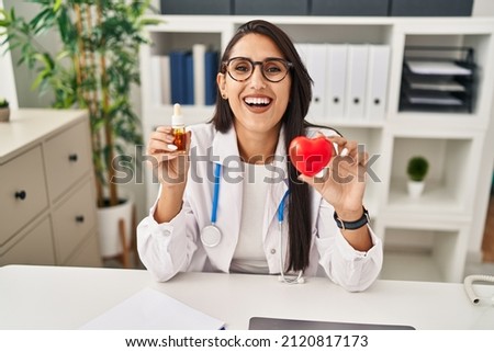 Young hispanic doctor woman holding heart and cbd oil smiling and laughing hard out loud because funny crazy joke. 
