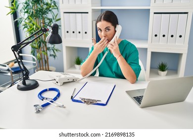 Young Hispanic Doctor Woman Speaking On The Phone At The Clinic Covering Mouth With Hand, Shocked And Afraid For Mistake. Surprised Expression 