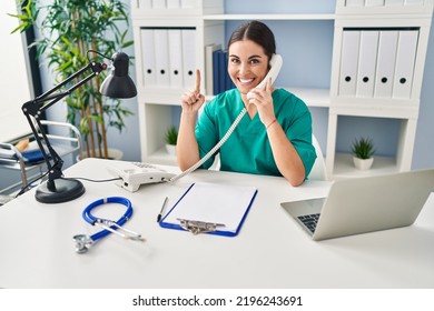 Young Hispanic Doctor Woman Speaking On The Phone At The Clinic Smiling With An Idea Or Question Pointing Finger With Happy Face, Number One 