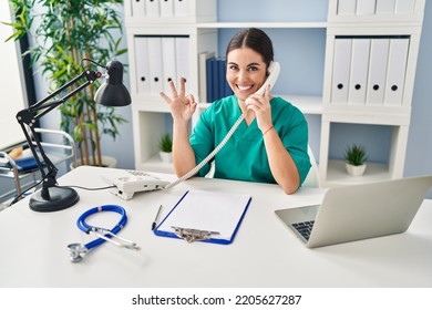 Young Hispanic Doctor Woman Speaking On The Phone At The Clinic Doing Ok Sign With Fingers, Smiling Friendly Gesturing Excellent Symbol 