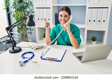 Young Hispanic Doctor Woman Speaking On The Phone At The Clinic Smiling Happy Pointing With Hand And Finger To The Side 