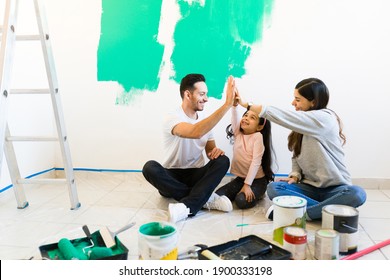 Young Hispanic Family Sitting On The Floor And Doing A High Five. Happy Mom, Dad And Little Girl Taking A Break From Painting And Remodeling The Living Room 