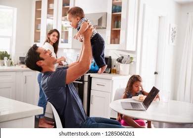 Young Hispanic Family In Their Kitchen, Dad Lifting Baby In The Air, Mum Cooking At Hob, Close Up