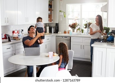 Young Hispanic Family In Their Kitchen, Dad Holding Baby In The Air, Mum Cooking At The Hob