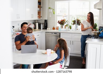 Young Hispanic Family In Their Kitchen, Dad Holding Baby, Mum Cooking At The Hob