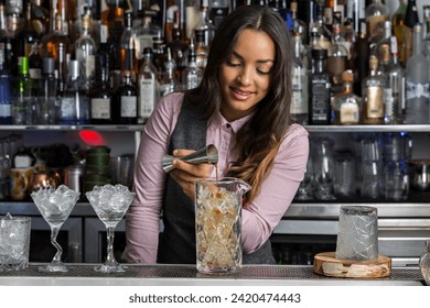 Young Hispanic female bartender adding alcohol from stainless jigger into mixing glass with ice cubes while preparing cocktail at bar counter