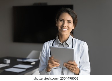 Young Hispanic female physician in coat posing for cam, holds digital tablet, reviewing patient records, collaborating with colleagues, check anamnesis. Healthcare professional using modern technology - Powered by Shutterstock