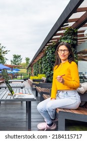 Young Hispanic Female Student Wearing A Yellow Sweater Looking Directly At The Camera In Front Of Her Laptop Sitting At A Table Outside