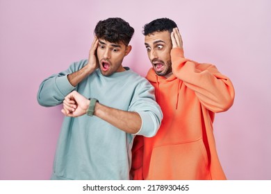 Young Hispanic Gay Couple Standing Over Pink Background Looking At The Watch Time Worried, Afraid Of Getting Late 