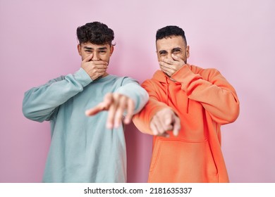 Young Hispanic Gay Couple Standing Over Pink Background Laughing At You, Pointing Finger To The Camera With Hand Over Mouth, Shame Expression 