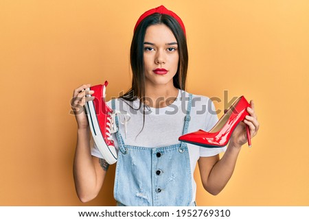 Young hispanic girl choosing high heel shoes and sneakers relaxed with serious expression on face. simple and natural looking at the camera. 