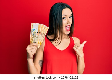 Young hispanic girl holding 500 philippine peso banknotes pointing thumb up to the side smiling happy with open mouth 