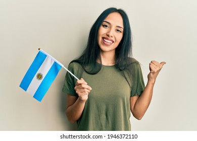 Young hispanic girl holding argentina flag pointing thumb up to the side smiling happy with open mouth 