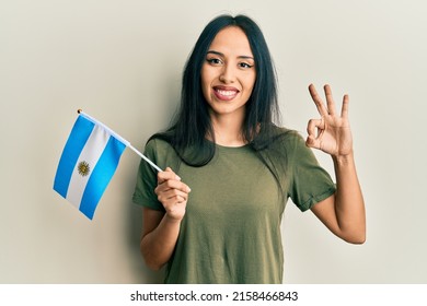 Young hispanic girl holding argentina flag doing ok sign with fingers, smiling friendly gesturing excellent symbol 