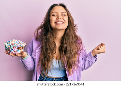 Young Hispanic Girl Holding Bowl Of Sugar Candy Screaming Proud, Celebrating Victory And Success Very Excited With Raised Arm 