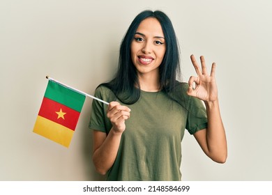 Young hispanic girl holding cameroon flag doing ok sign with fingers, smiling friendly gesturing excellent symbol 