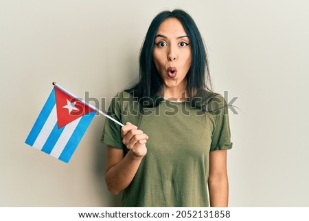 Young hispanic girl holding cuba flag scared and amazed with open mouth for surprise, disbelief face 
