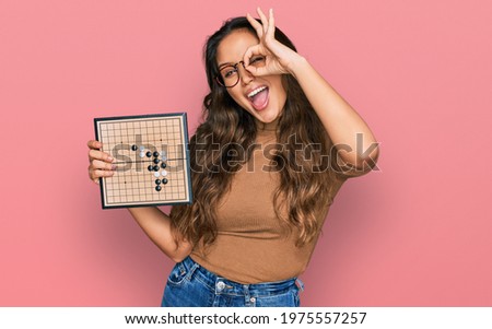 Young hispanic girl holding drauhgts smiling happy doing ok sign with hand on eye looking through fingers 