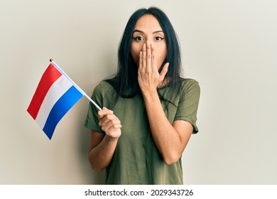 Young hispanic girl holding holland flag covering mouth with hand, shocked and afraid for mistake. surprised expression 