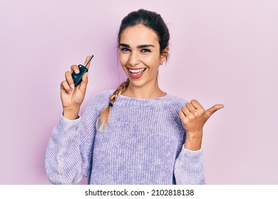 Young hispanic girl holding key of new car pointing thumb up to the side smiling happy with open mouth 