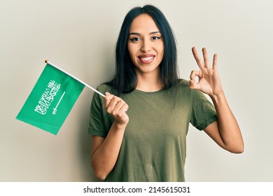 Young Hispanic Girl Holding Kingdom Of Saudi Arabia Flag Doing Ok Sign With Fingers, Smiling Friendly Gesturing Excellent Symbol 