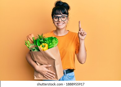 Young hispanic girl holding paper bag with bread and groceries smiling with an idea or question pointing finger with happy face, number one 