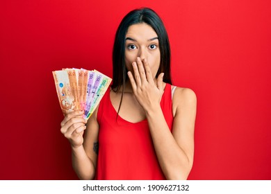 Young hispanic girl holding philippine peso banknotes covering mouth with hand, shocked and afraid for mistake. surprised expression 