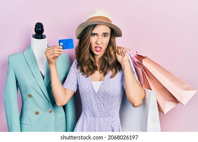 Young Hispanic Girl Holding Shopping Bags And Credit Card Clueless And Confused Expression. Doubt Concept. 